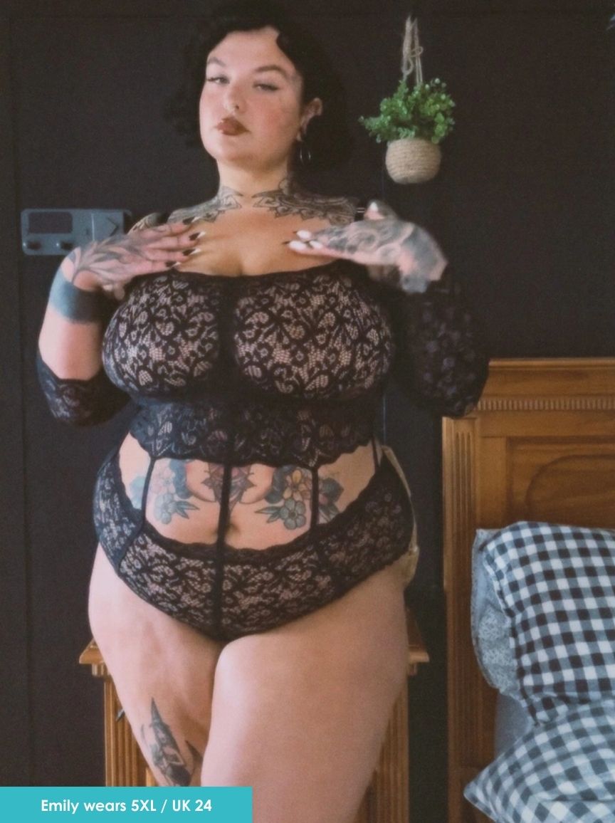 Person wearing a black lace bodysuit in a room with a plant and wooden furniture.